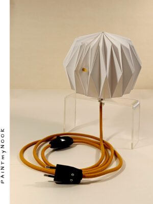 A white paper lamp folded using origami techniques, attached to a transparent plexiglass prop, with a mustard yellow textile cable