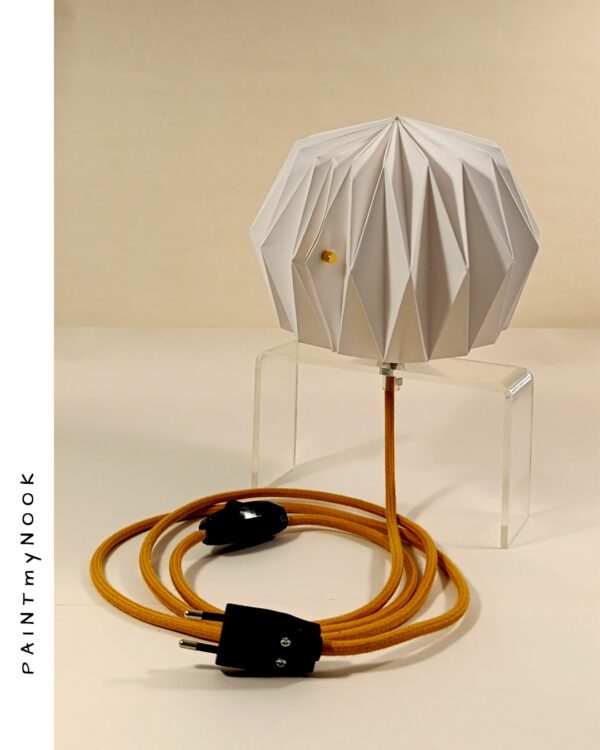 A white paper lamp folded using origami techniques, attached to a transparent plexiglass prop, with a mustard yellow textile cable