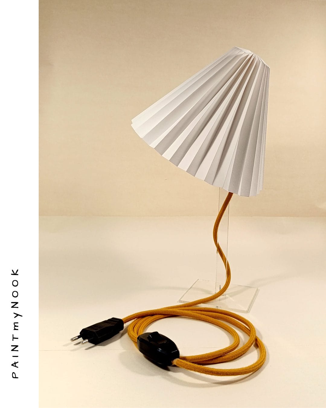 A table top white origami paper lamp with a mustard yellow textile cable on a transparent plexiglas prop.